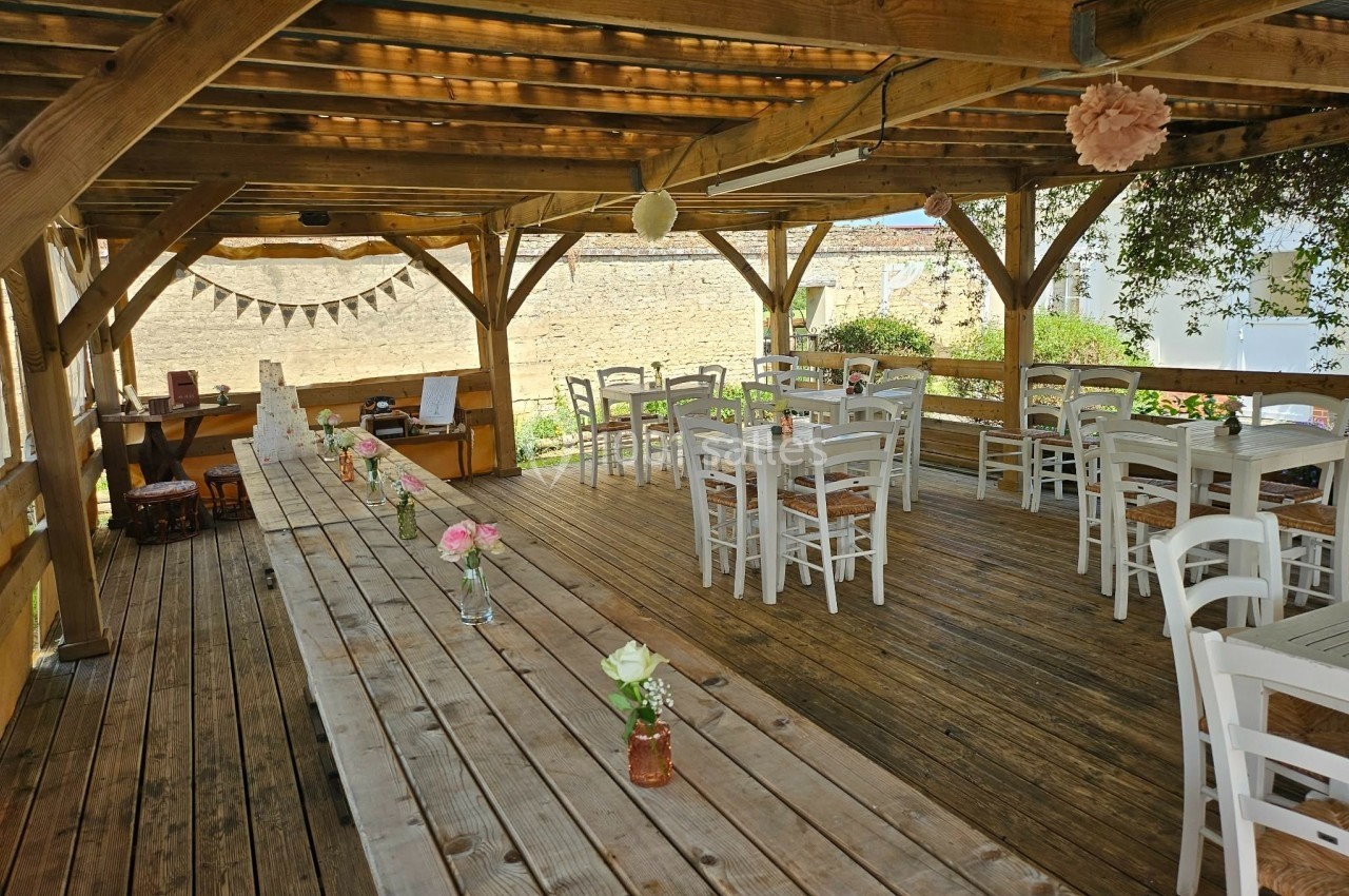 Terrasse en bois couverte avec tables et chaises blanches, décorée de fleurs et guirlandes dans un cadre champêtre.
