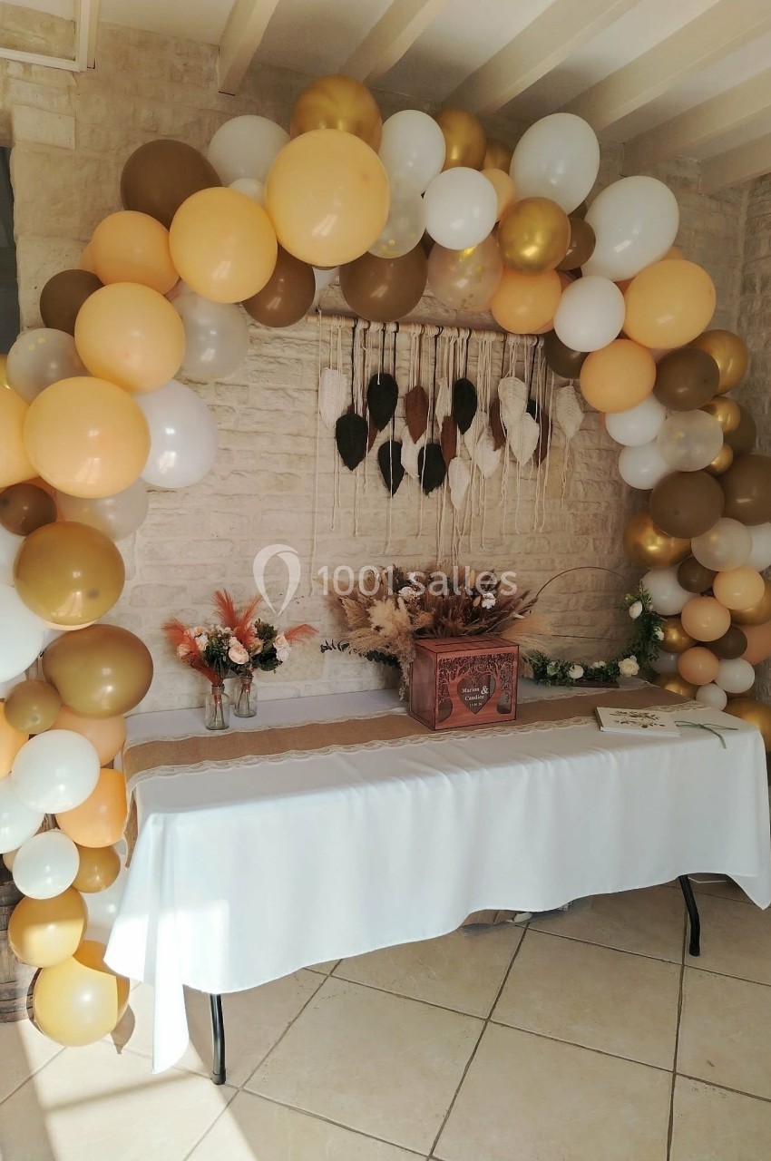 Arche de ballons blancs, dorés et beige décorant une table avec fleurs séchées et caisse en bois dans une pièce lumineuse.