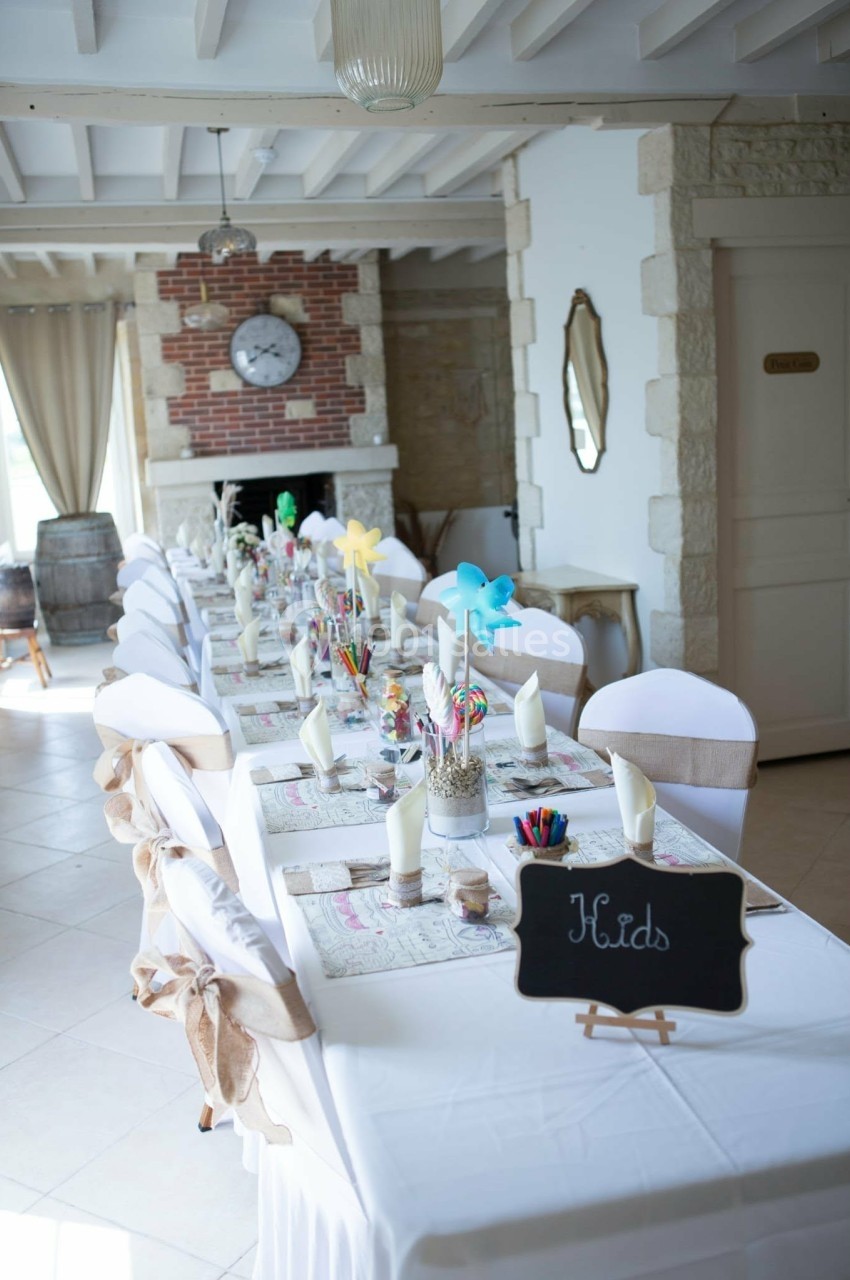 Table décorée pour un événement, avec nappes blanches, serviettes pliées, décorations colorées et ardoise indiquant ’Kids’.