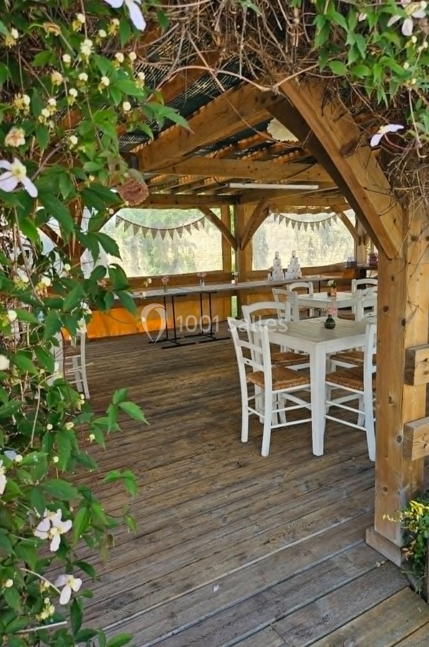 Terrasse en bois couverte avec table et chaises blanches, décorée de plantes grimpantes et guirlandes.