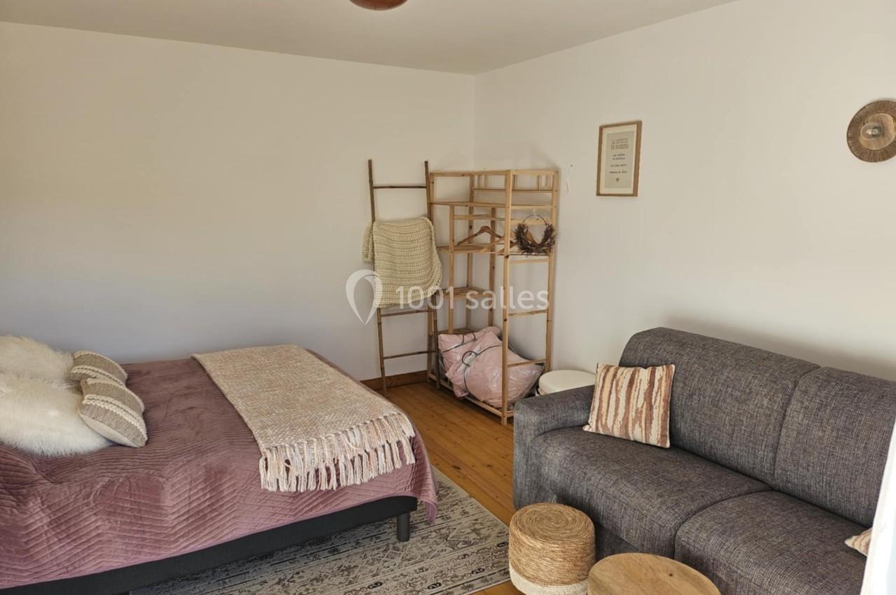 Chambre lumineuse avec lit double, canapé gris, étagère en bois, décor minimaliste et parquet au sol.