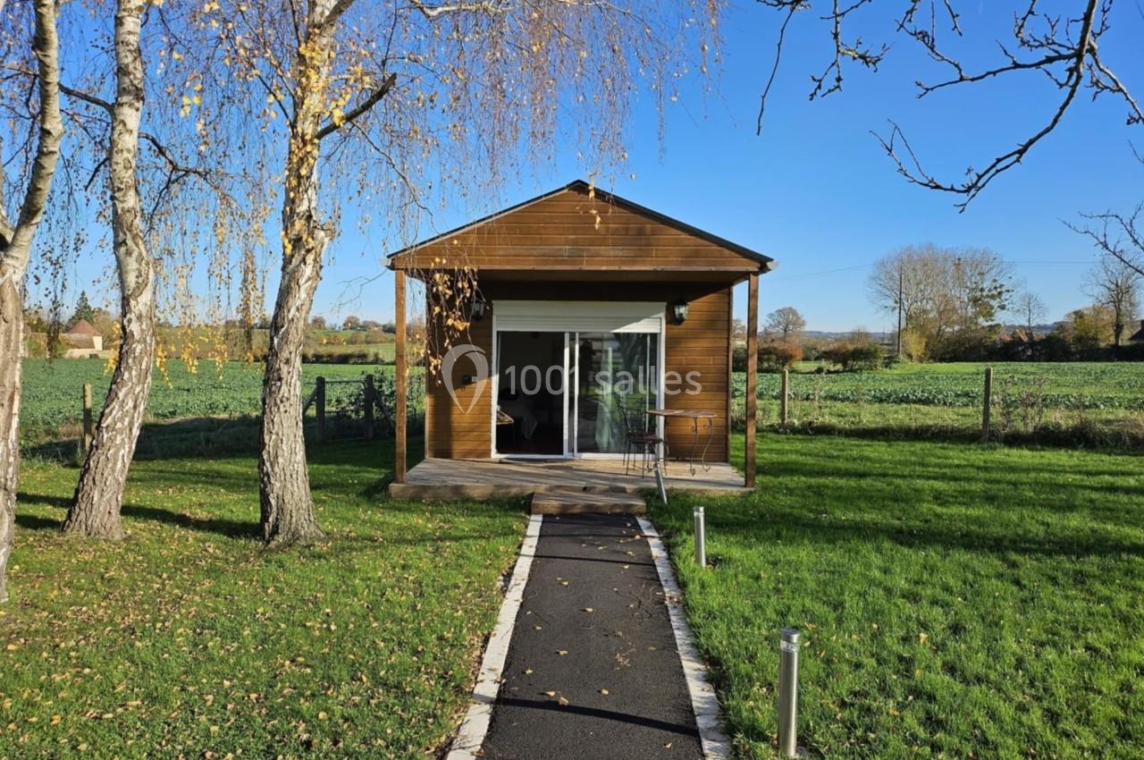 Petite maison en bois avec terrasse, entourée de pelouse et d'arbres, située dans un cadre champêtre.