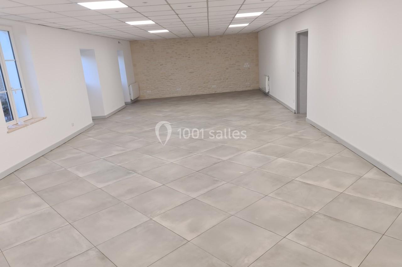 Salle vide avec sol en carrelage gris, murs blancs, plafond à dalles et une fenêtre laissant entrer la lumière naturelle.