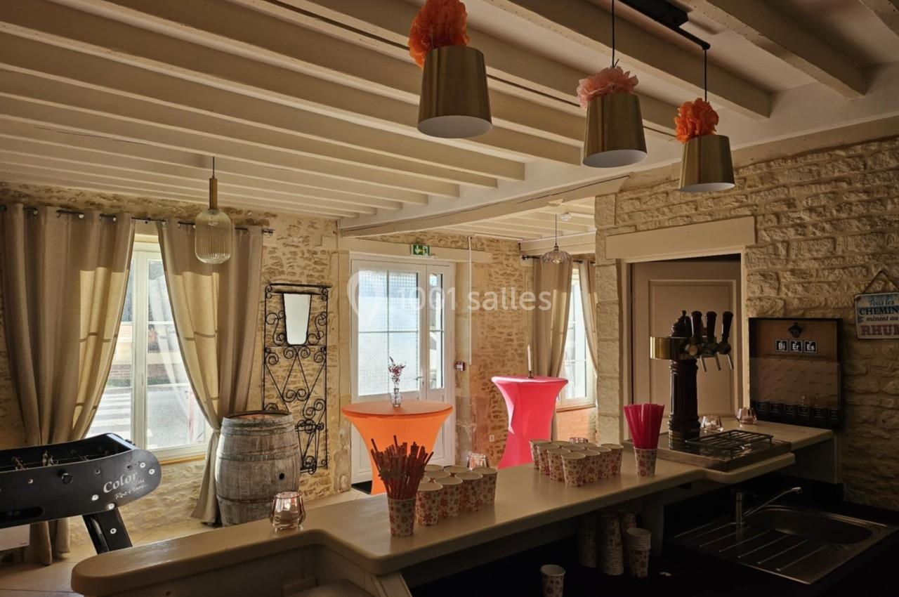 Salle lumineuse avec comptoir, baby-foot, tonneau décoratif et tables hautes colorées sous un plafond à poutres apparentes.