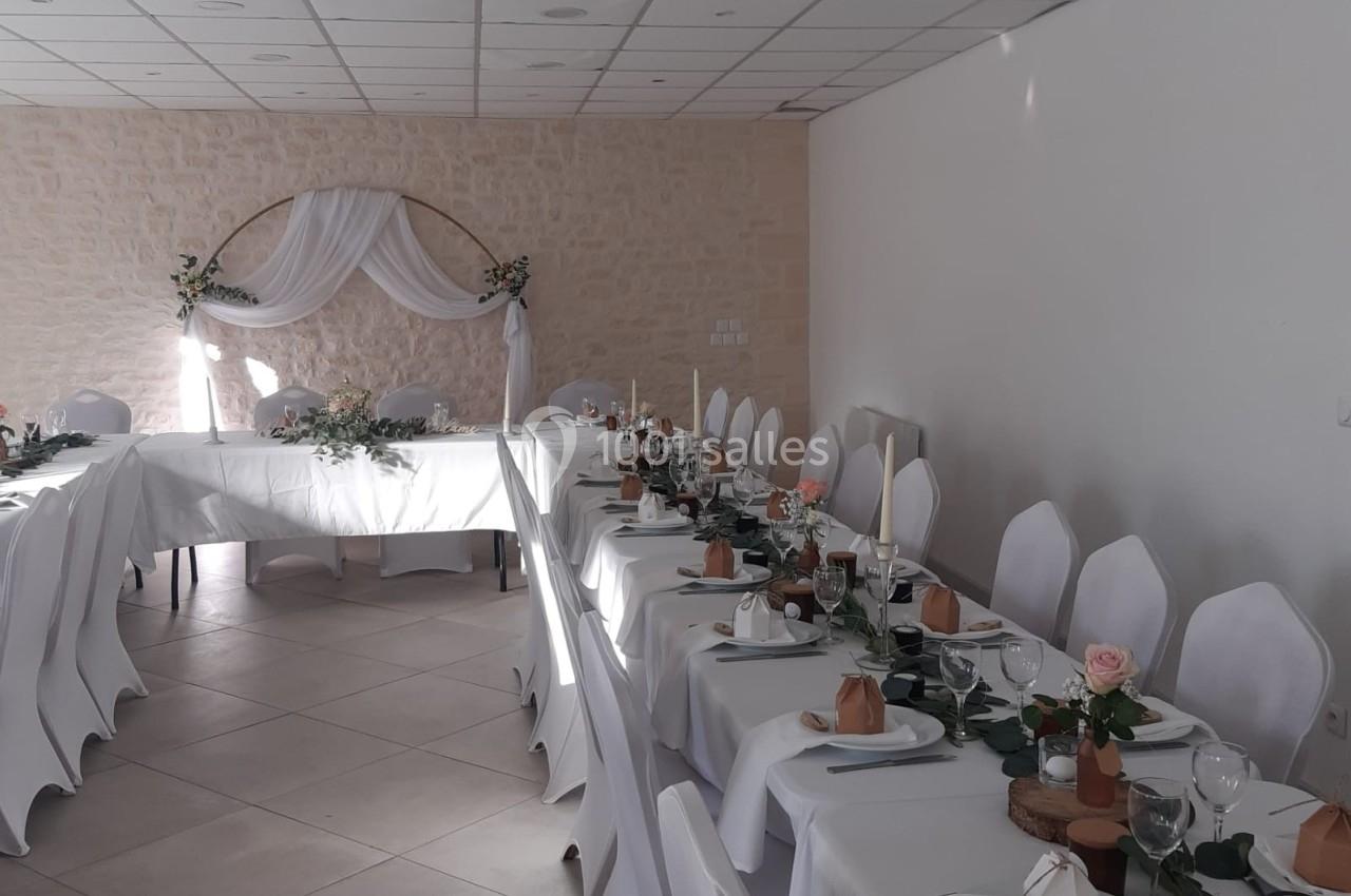 Salle de réception décorée avec des tables en U, nappes blanches, vaisselle élégante et arche florale en arrière-plan.