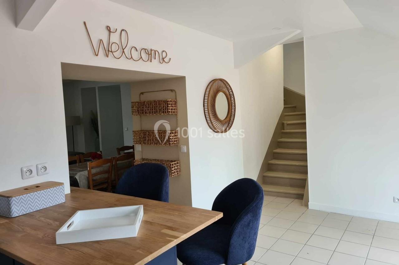 Cuisine ouverte avec comptoir en bois, chaises bleues, miroir rond, étagères murales et escalier en arrière-plan.