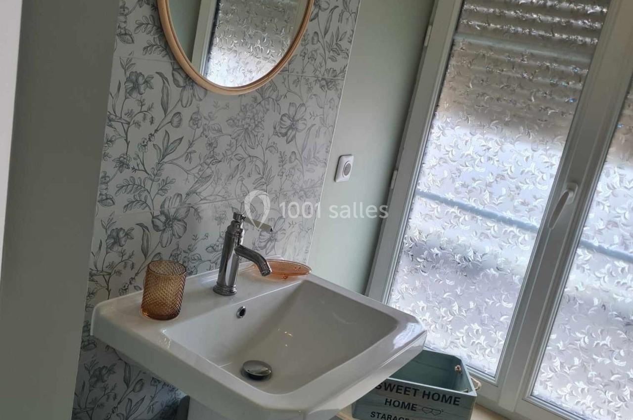 Lavabo blanc avec miroir rond, mur à motif floral, fenêtre avec stores baissés et étagère en bois.