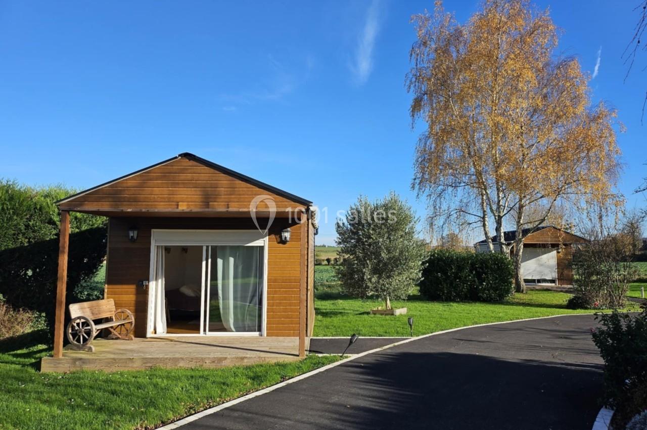 Chalet en bois avec terrasse, entouré de pelouse et d'arbres, sous un ciel bleu clair.