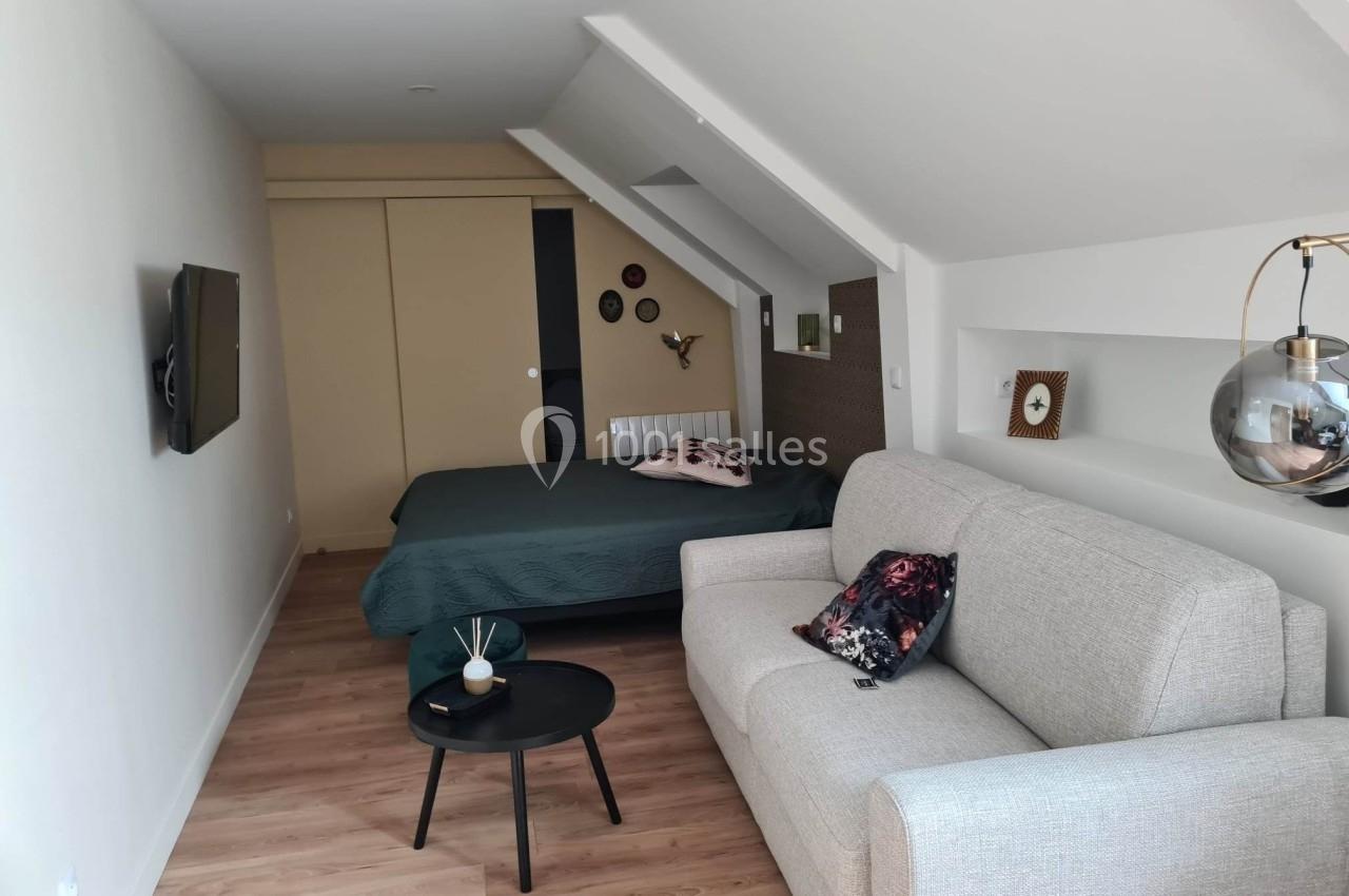 Studio mansardé avec lit double, canapé, table basse et décoration murale, éclairé par une lumière naturelle.