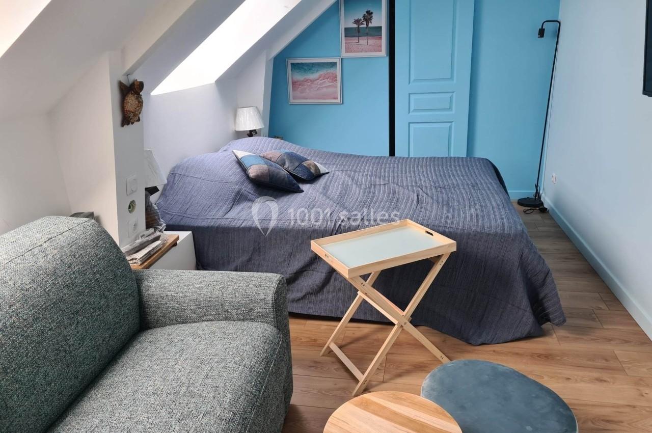 Chambre mansardée avec lit double, canapé, table pliante, pouf et décoration murale sur fond bleu.