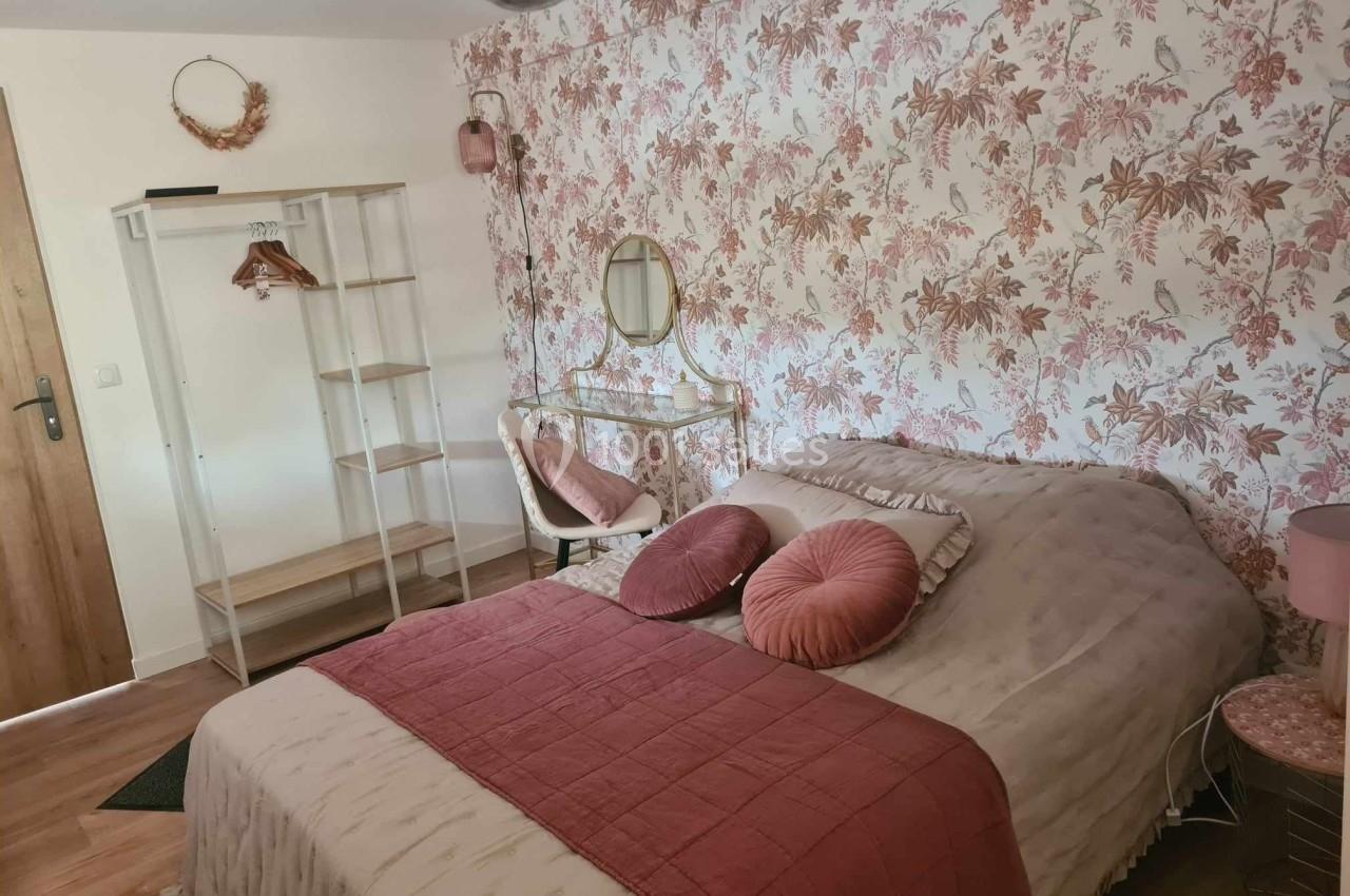 Chambre décorée avec papier peint floral, lit avec coussins roses, coiffeuse et portant en bois près d'une porte en bois.