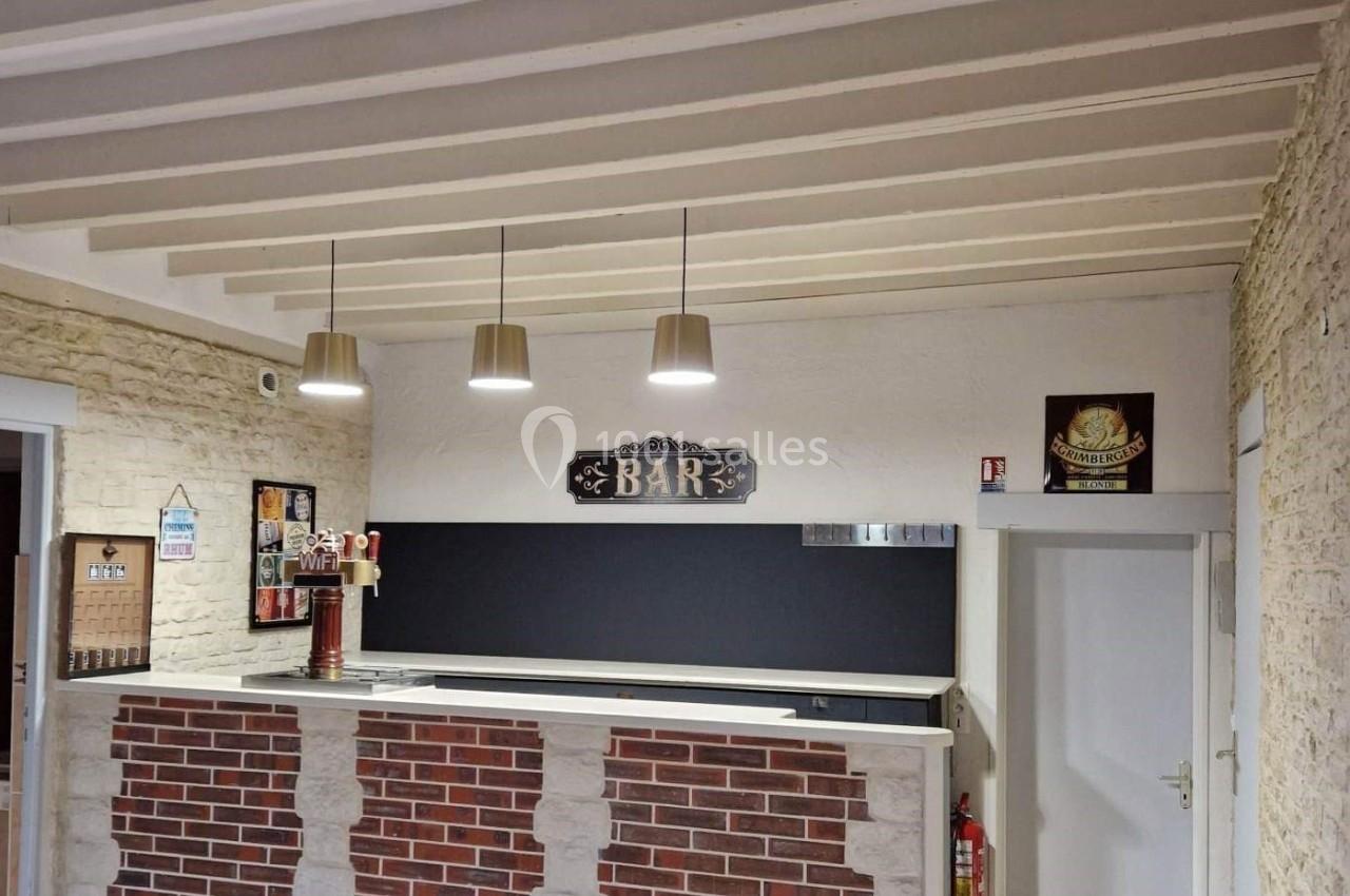 Comptoir de bar en briques avec panneau ’BAR’, trois suspensions lumineuses et murs en pierre apparente.