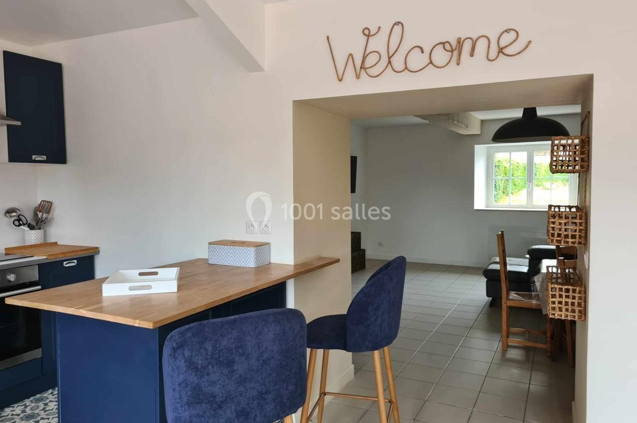 Cuisine ouverte avec comptoir en bois, tabourets bleus, et vue sur une salle à manger lumineuse, décorée d'un panneau…