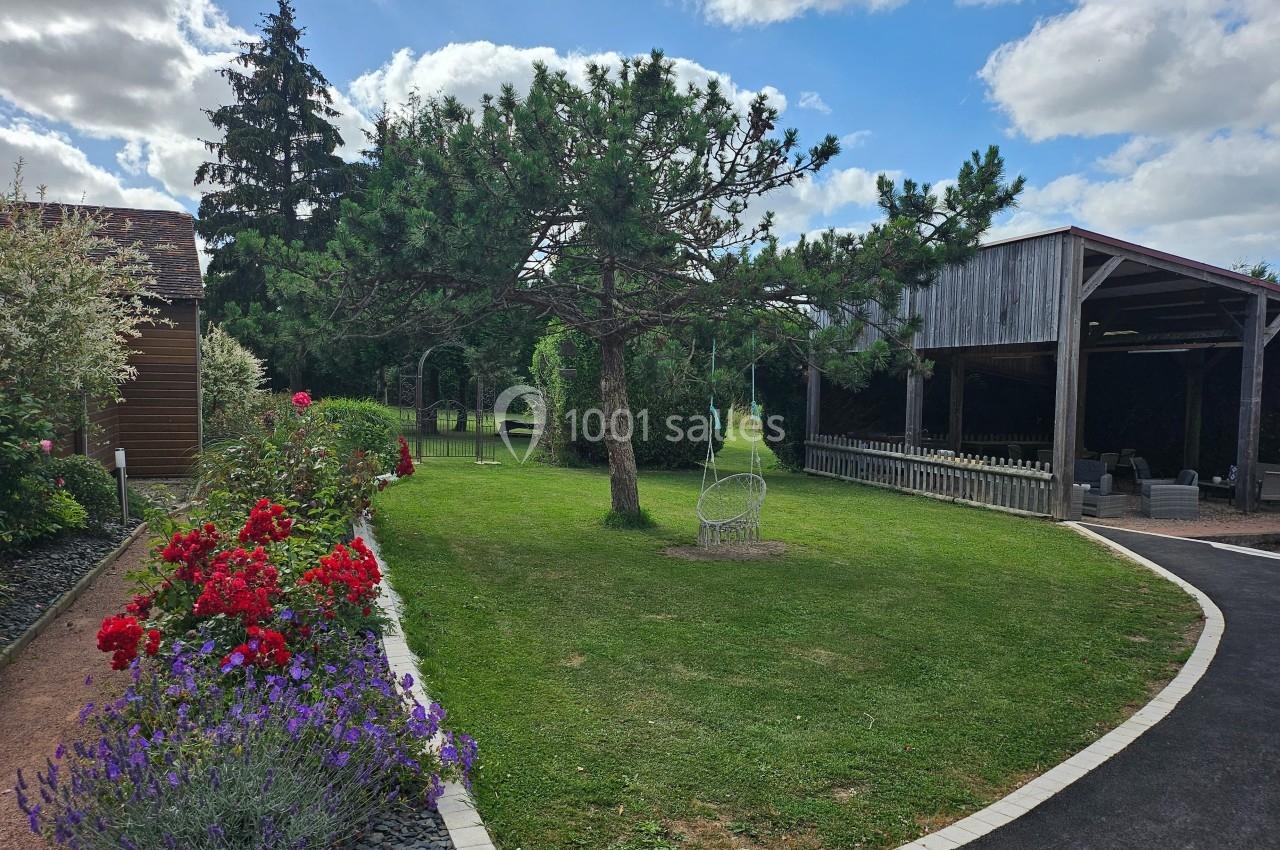 Jardin avec pelouse, fleurs colorées, arbre central, fauteuil suspendu et abri en bois sous un ciel partiellement nuageux.
