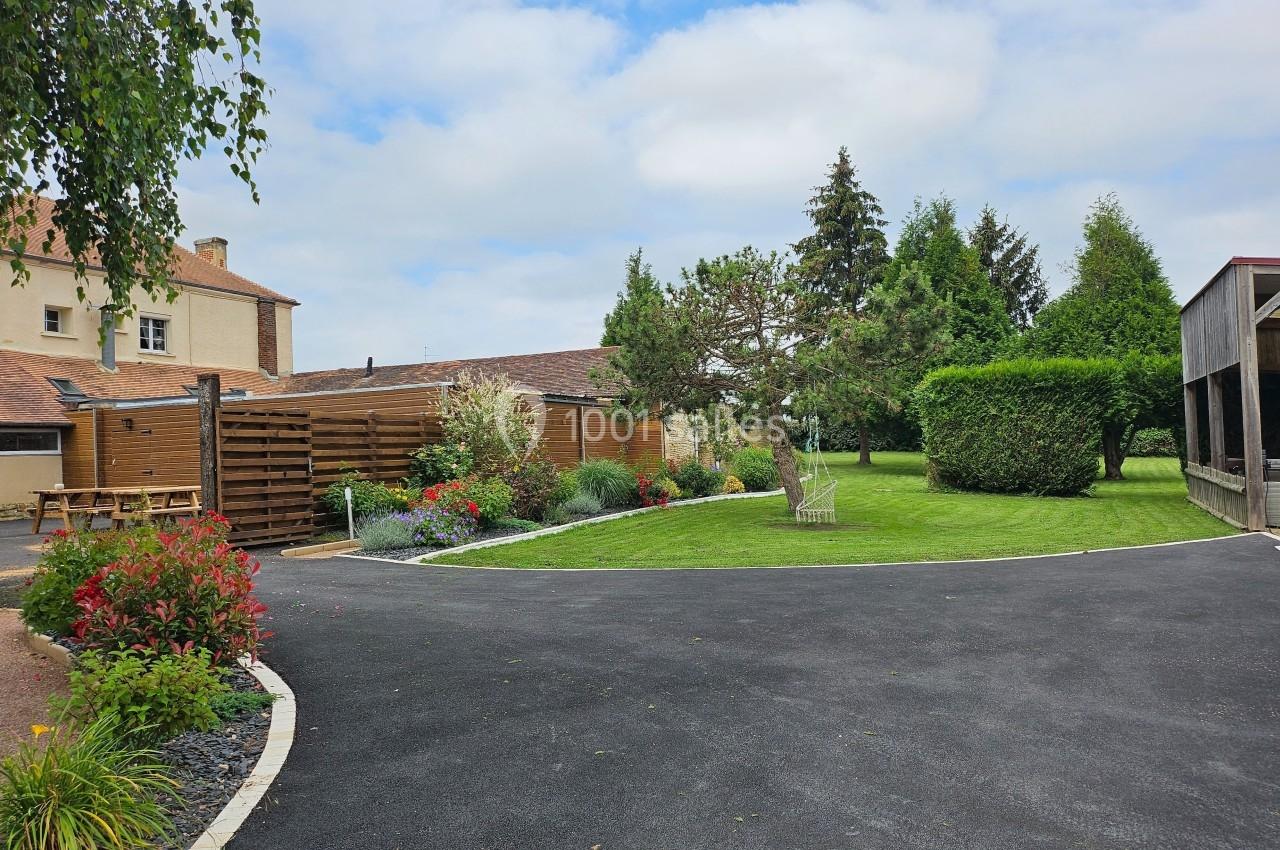 Cour avec allée asphaltée, pelouse, arbres, massifs de fleurs et bâtiments en arrière-plan sous un ciel partiellement…