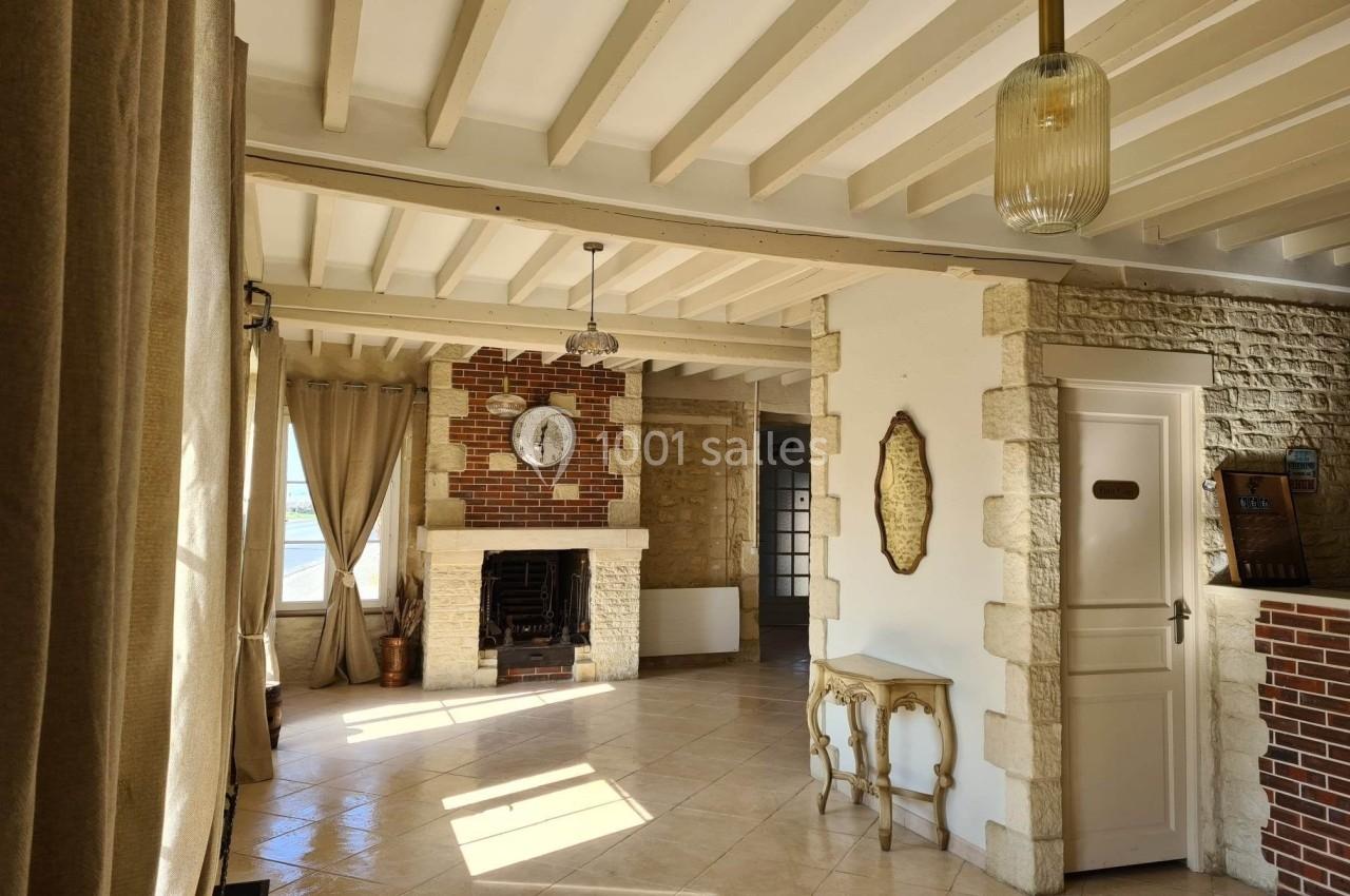 Salon lumineux avec murs en pierre et briques, cheminée centrale, poutres apparentes et grandes fenêtres avec rideaux.