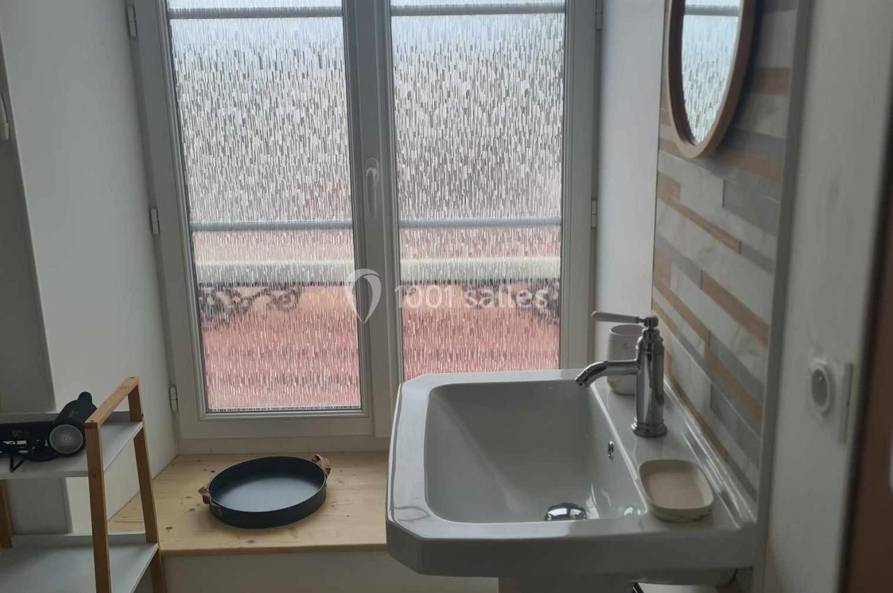 Lavabo blanc devant une fenêtre avec verre dépoli, étagère en bois à gauche et miroir rond au mur.
