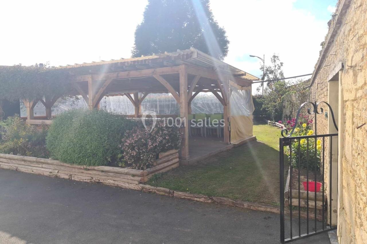 Abri en bois avec table et chaises, entouré de plantes, situé dans une cour avec un portail ouvert.