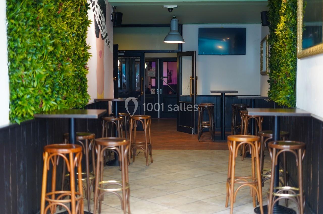 Intérieur d'un café avec des tabourets en bois, des tables hautes et des murs ornés de plantes vertes.