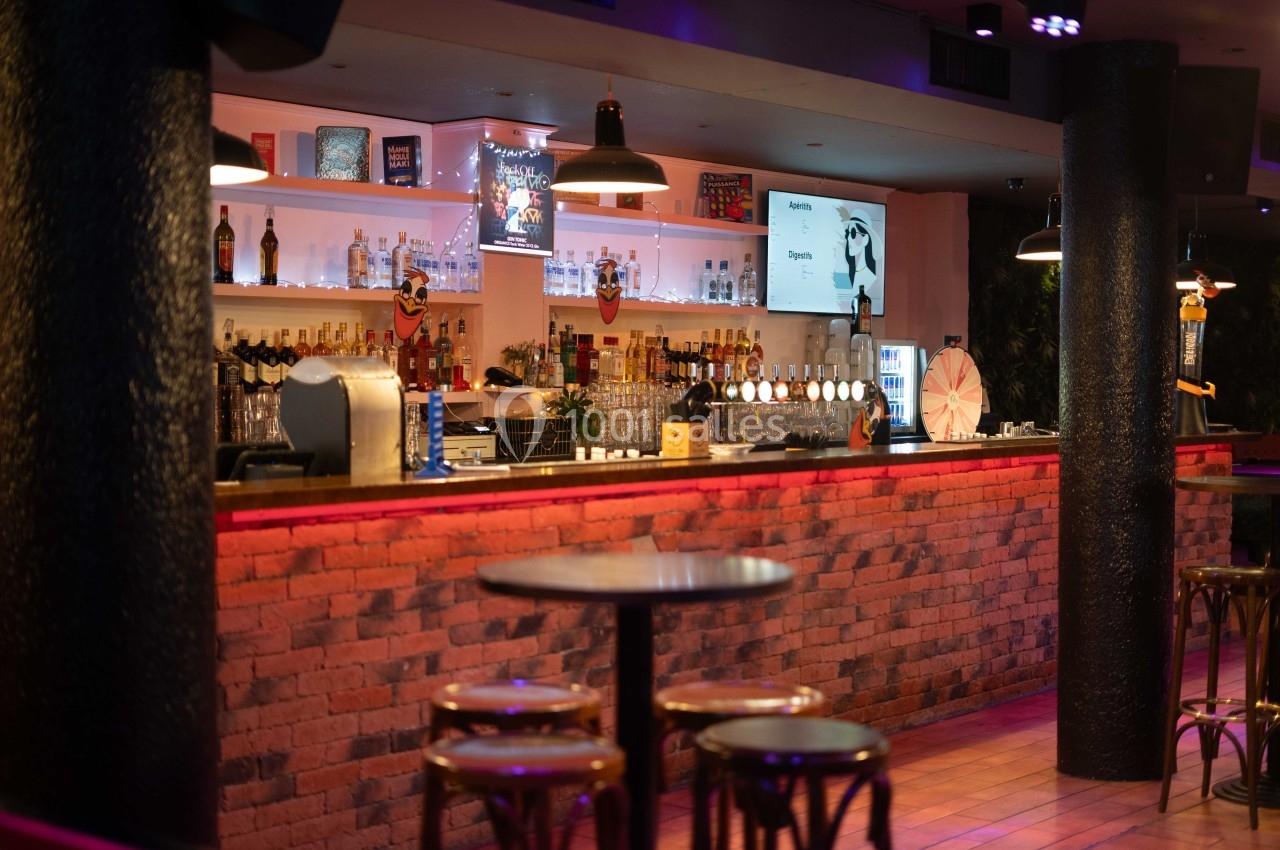 Bar avec comptoir en briques rouges, étagères éclairées exposant des bouteilles, et tables hautes au premier plan.