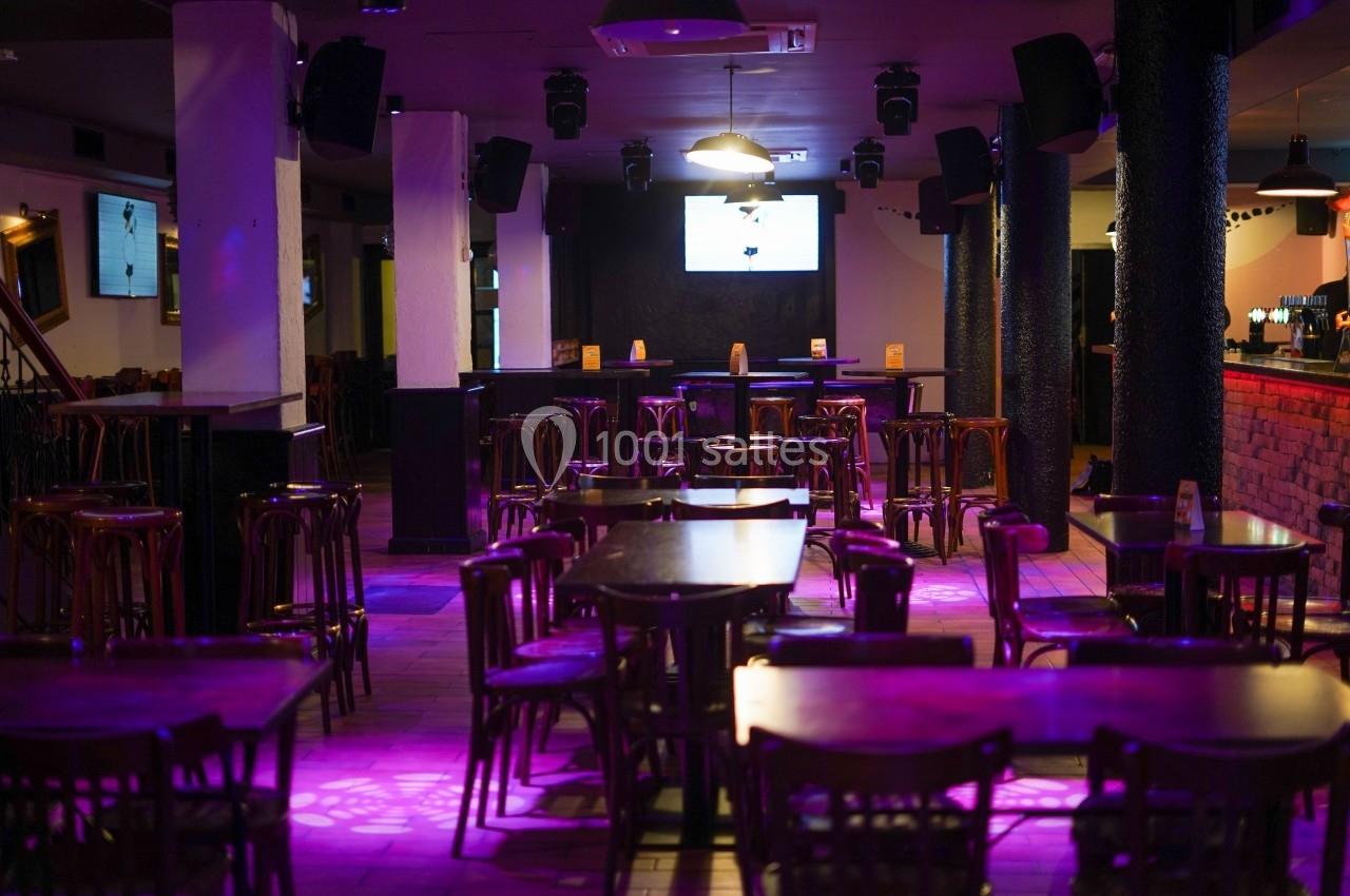 Salle de bar avec tables et chaises en bois, éclairage tamisé violet et écran suspendu au fond.