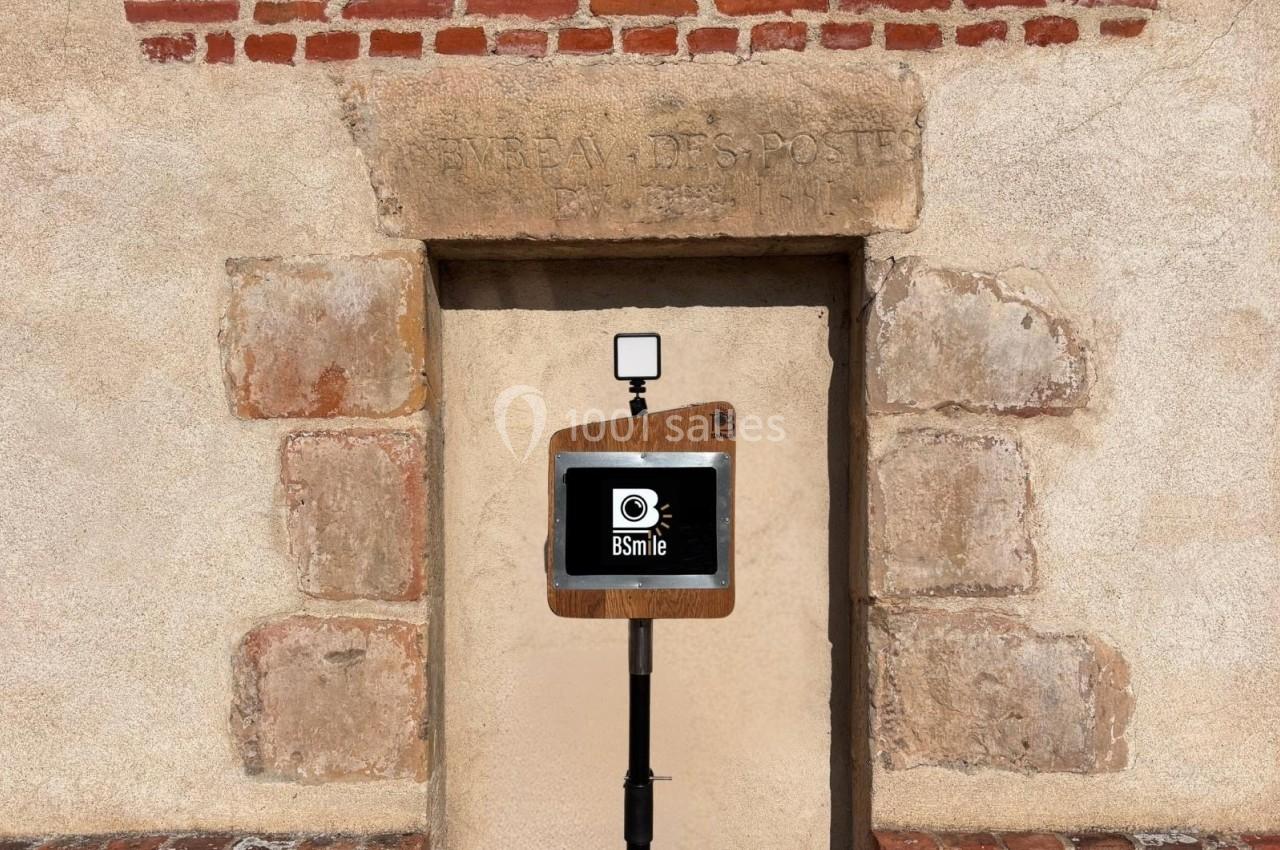 Borne photo en bois avec écran et flash, placée devant une porte en pierre encadrée de briques.