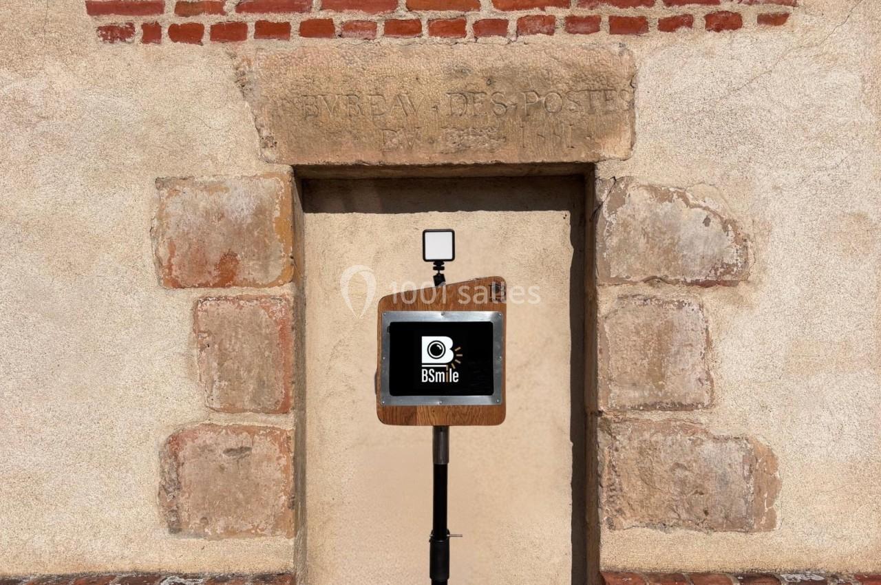 Borne photo en bois avec écran et flash, installée devant une porte en pierre et un mur en briques.