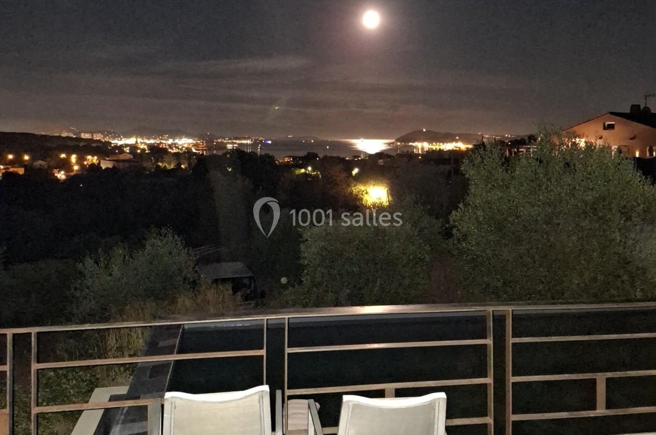 Vue nocturne depuis une terrasse avec des chaises, montrant une pleine lune éclairant une ville et un plan d'eau au loin.
