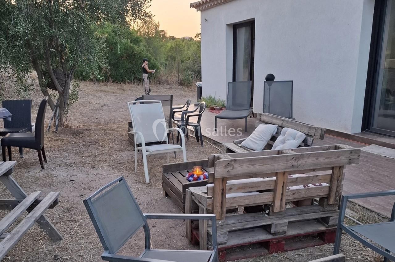 Terrasse extérieure avec mobilier en bois et en plastique, entourée d'arbres, une personne en arrière-plan.