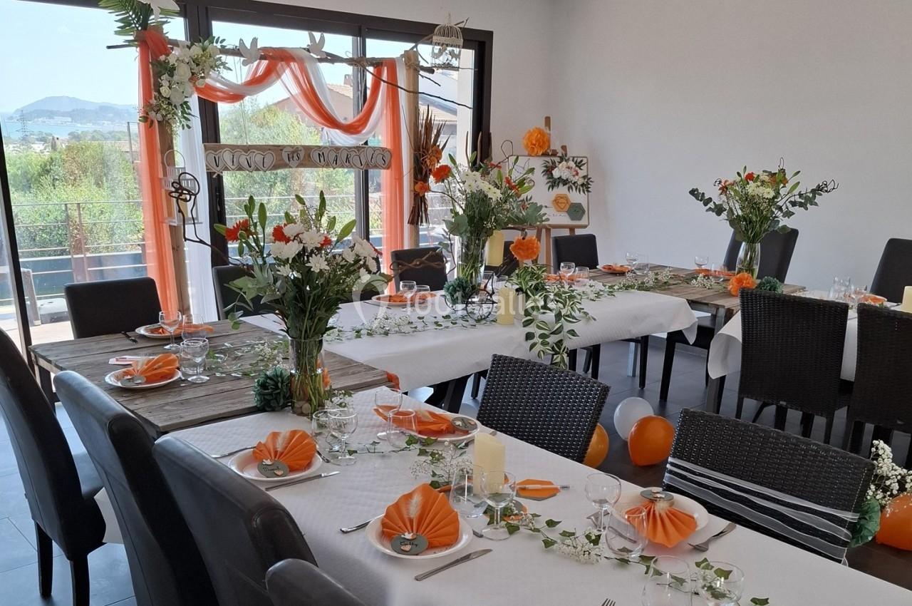 Salle décorée pour un événement avec tables dressées, nappes blanches, serviettes orange et arrangements floraux.