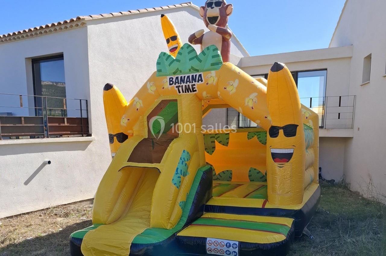 Structure gonflable jaune en forme de bananes avec toboggan, installée dans un jardin devant une maison.