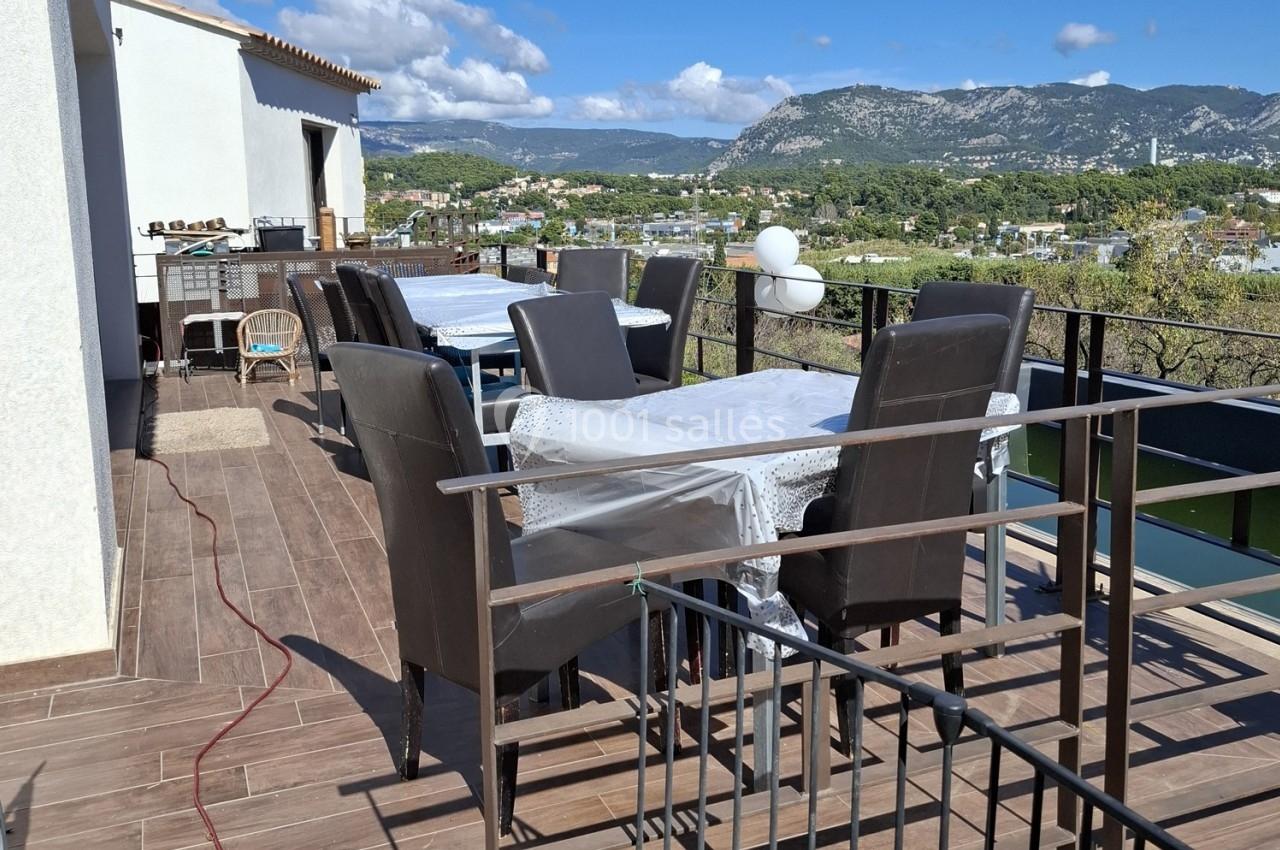 Terrasse avec tables et chaises noires, vue sur un paysage vallonné sous un ciel partiellement nuageux.