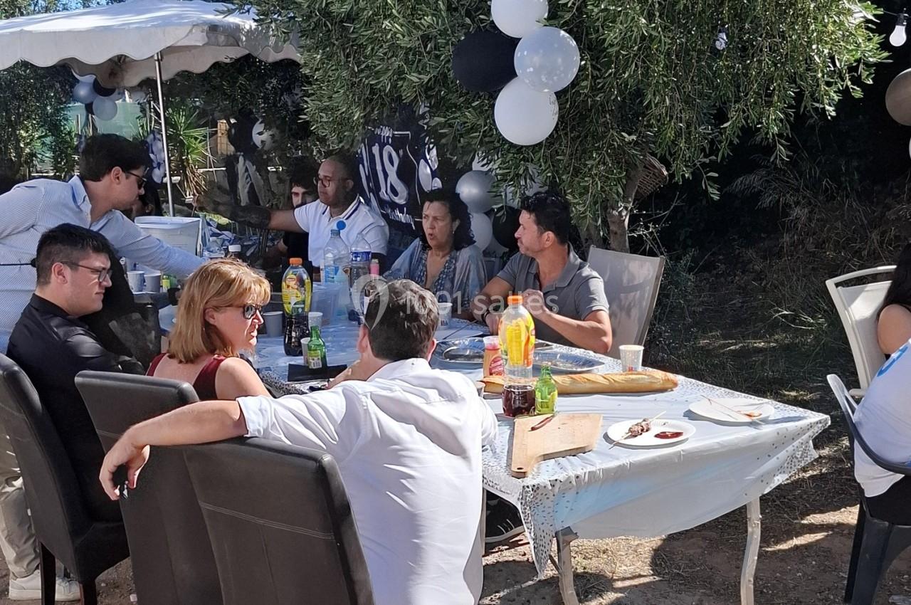 Des personnes assises autour d'une table en extérieur, décorée pour une fête, avec nourriture et boissons.