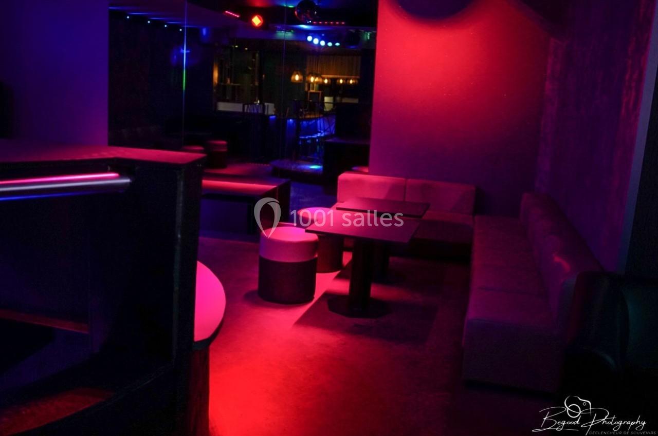 Salle de club avec éclairage tamisé rouge et violet, banquettes, tables basses et ambiance nocturne.