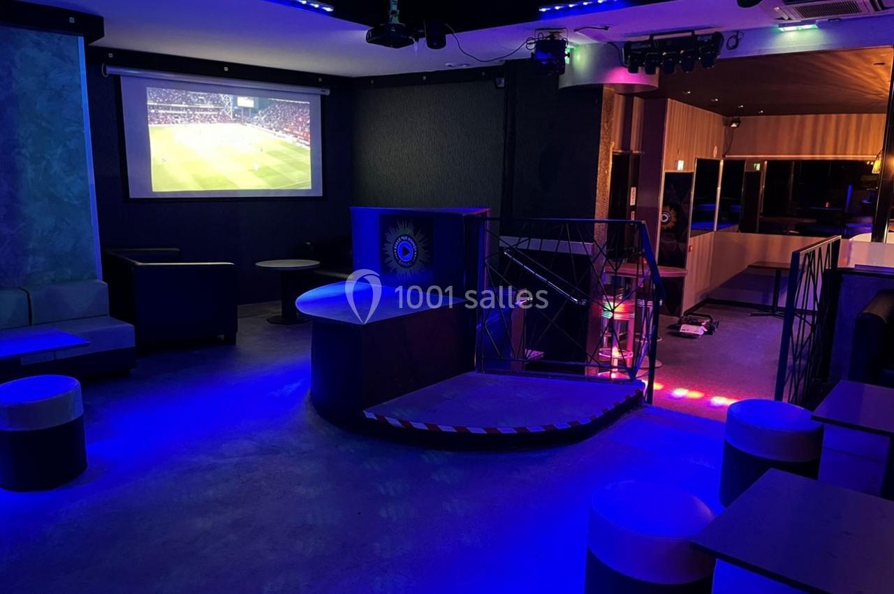 Salle sombre avec éclairage bleu, écran diffusant un match de football, tables et sièges disposés autour.