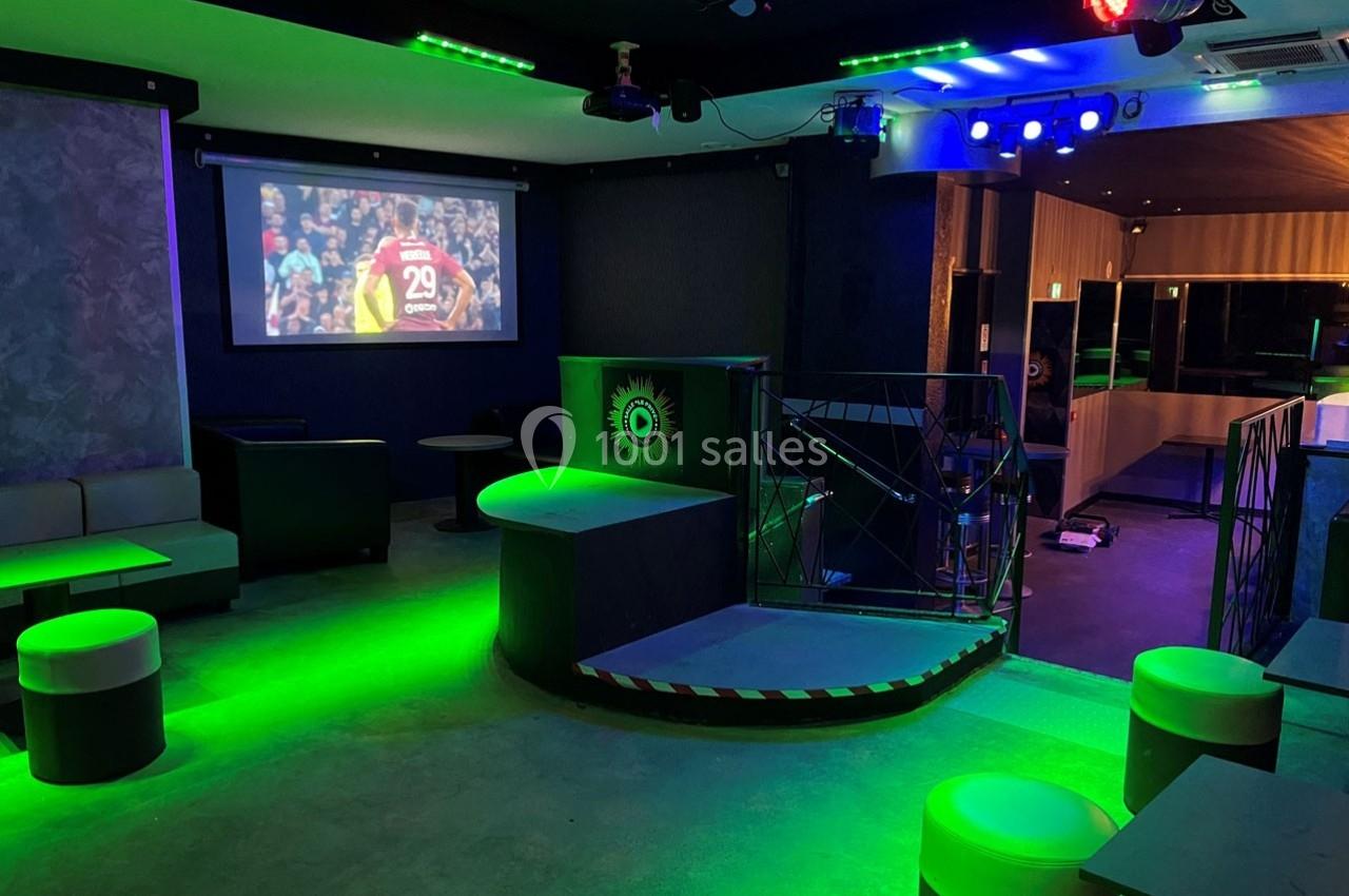 Salle de bar avec éclairage vert, écran diffusant un match de football et mobilier moderne.