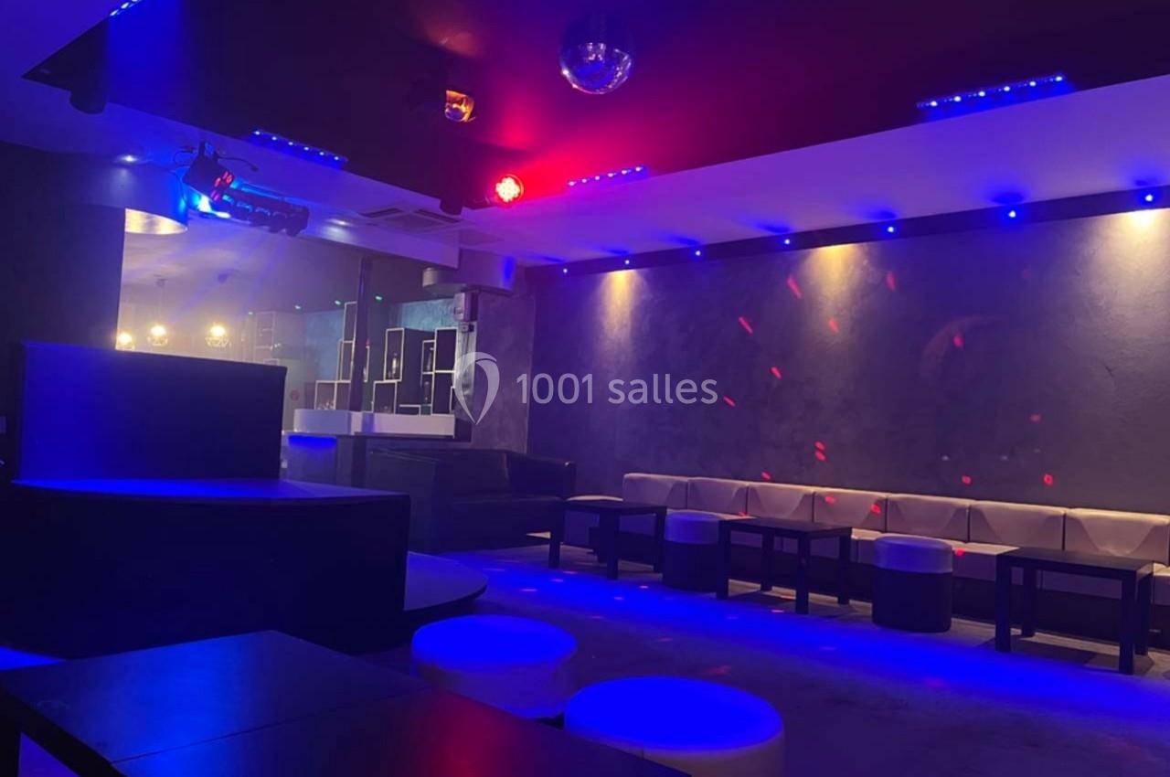 Salle de club avec éclairage tamisé bleu et rouge, banquettes, tables basses et piste de danse vide.