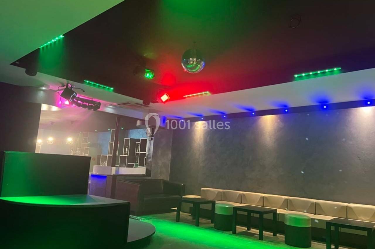 Salle de club avec éclairage coloré, banquettes, tables basses et une boule disco suspendue au plafond.