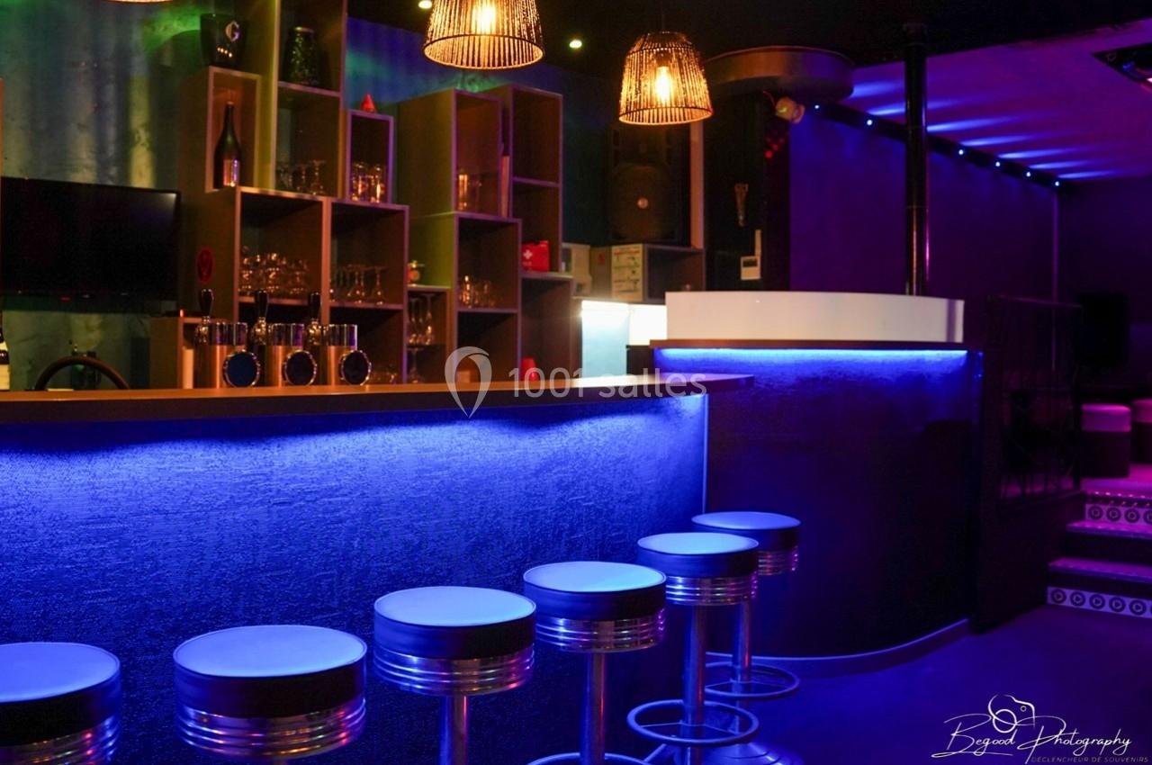 Bar avec comptoir éclairé en bleu, tabourets métalliques alignés et étagères contenant des verres en arrière-plan.