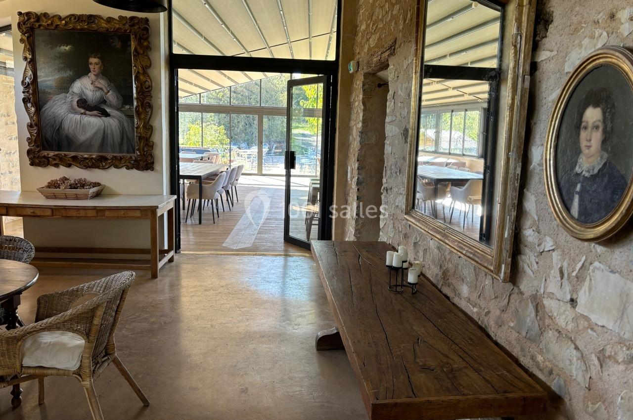 Salle lumineuse avec murs en pierre, grandes fenêtres, tables en bois, et portraits encadrés accrochés aux murs.