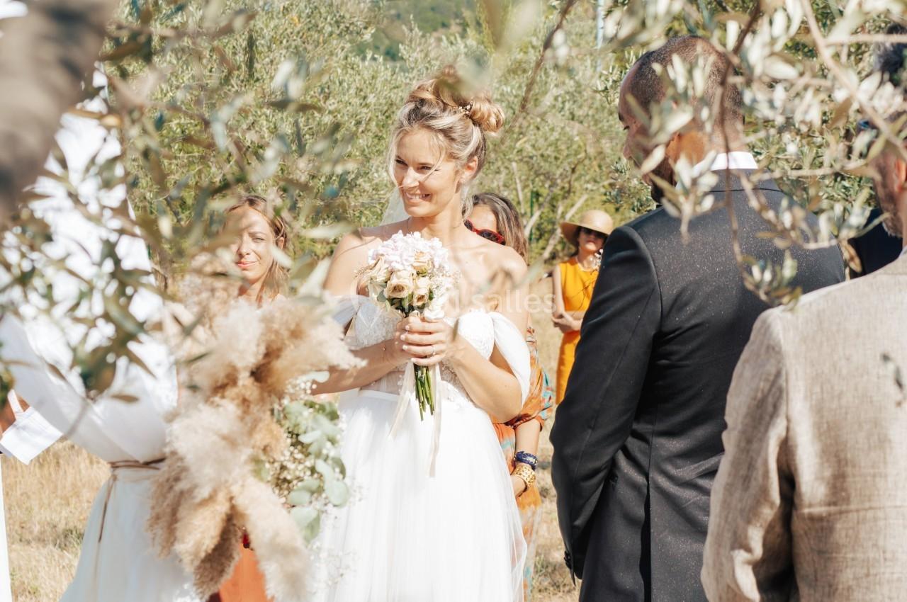 Une mariée en robe blanche tient un bouquet de fleurs lors d'une cérémonie en plein air entourée d'invités.