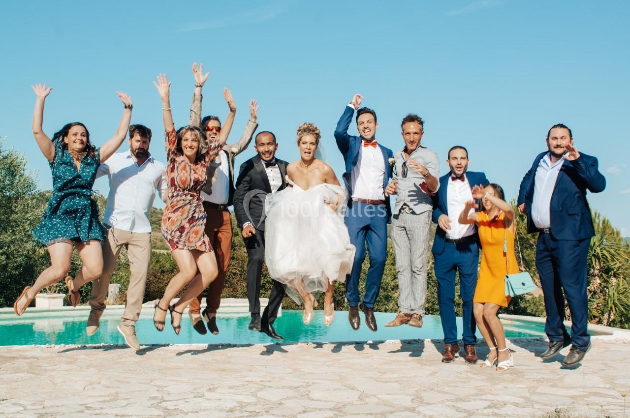 Un groupe de personnes, dont un couple en tenue de mariage, saute joyeusement devant une piscine en plein air.