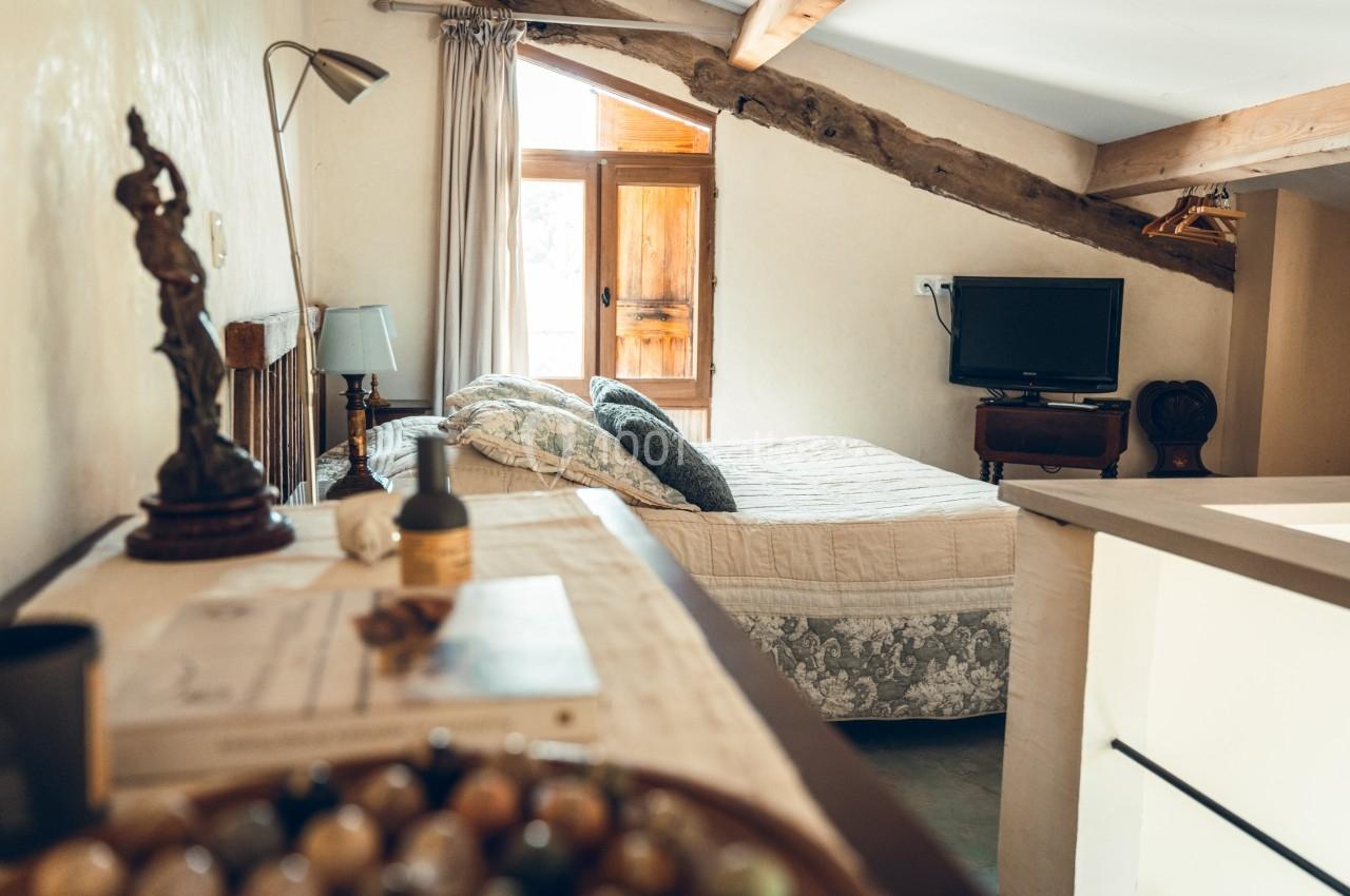 Chambre lumineuse avec lit double, télévision, poutres apparentes et vue sur une porte-fenêtre en bois.