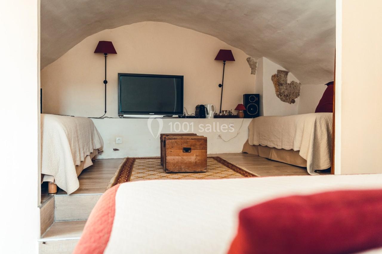 Chambre mansardée avec deux lits simples, une télévision, des lampes murales et un coffre en bois au centre.