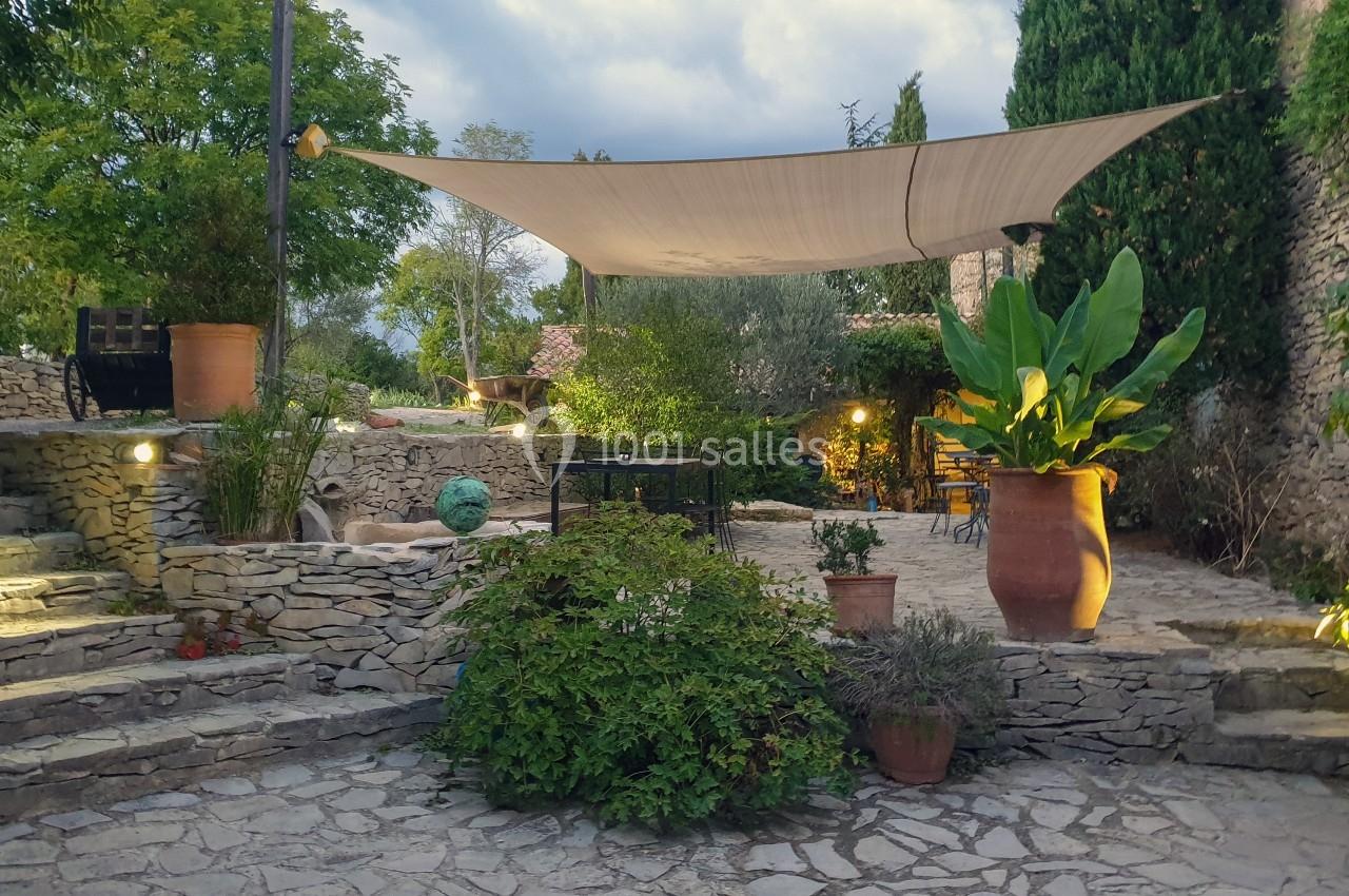 Cour en pierre avec des plantes en pot, un voile d'ombrage tendu et un éclairage doux en soirée.
