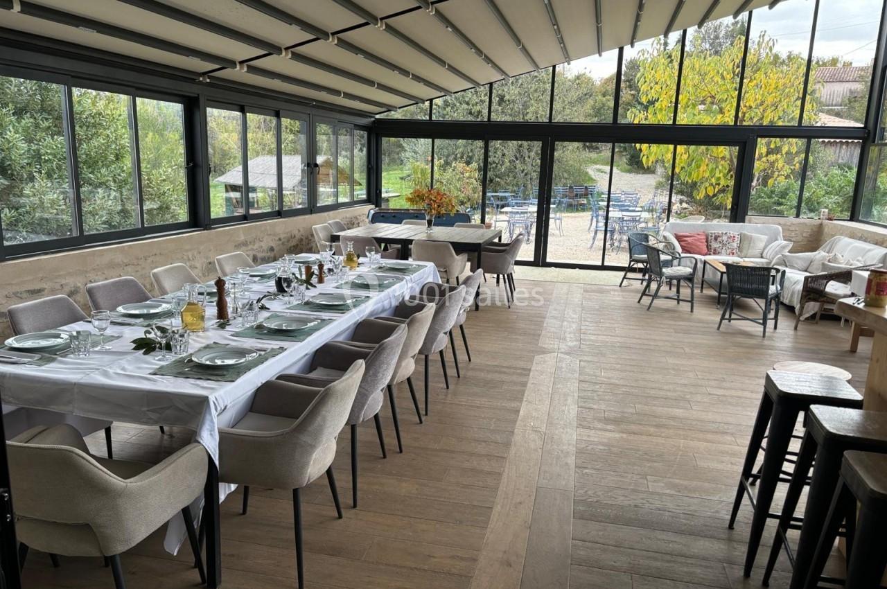Salle lumineuse avec de grandes baies vitrées, tables dressées pour un repas, chaises confortables et vue sur un jardin.