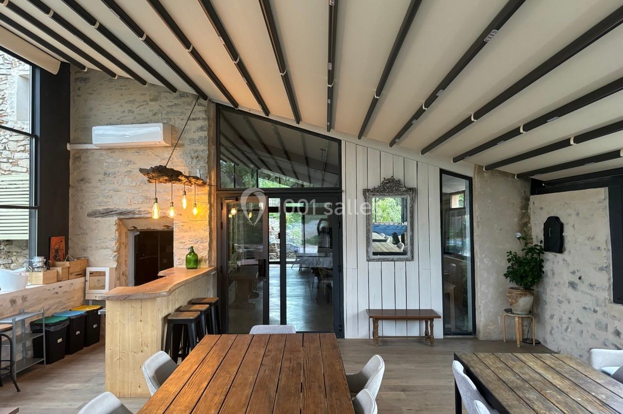 Salle lumineuse avec murs en pierre, tables en bois, bar avec suspensions, et grandes baies vitrées donnant sur l'extérieur.