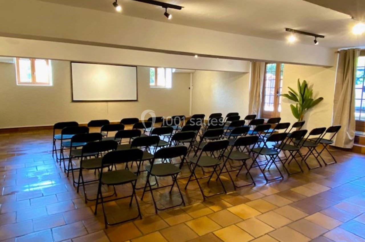 Salle de réunion avec rangées de chaises noires, un écran blanc, sol en carrelage et lumière naturelle.