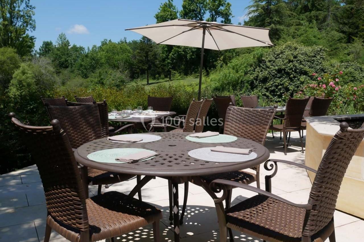 Terrasse extérieure avec tables en métal, chaises en osier et parasol, entourée de verdure sous un ciel bleu.