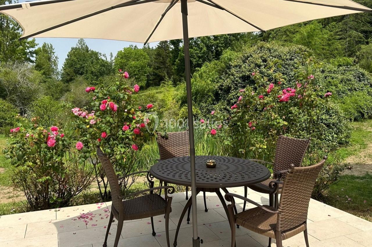 Table et chaises en métal sous un parasol beige, entourées de rosiers fleuris dans un jardin verdoyant.